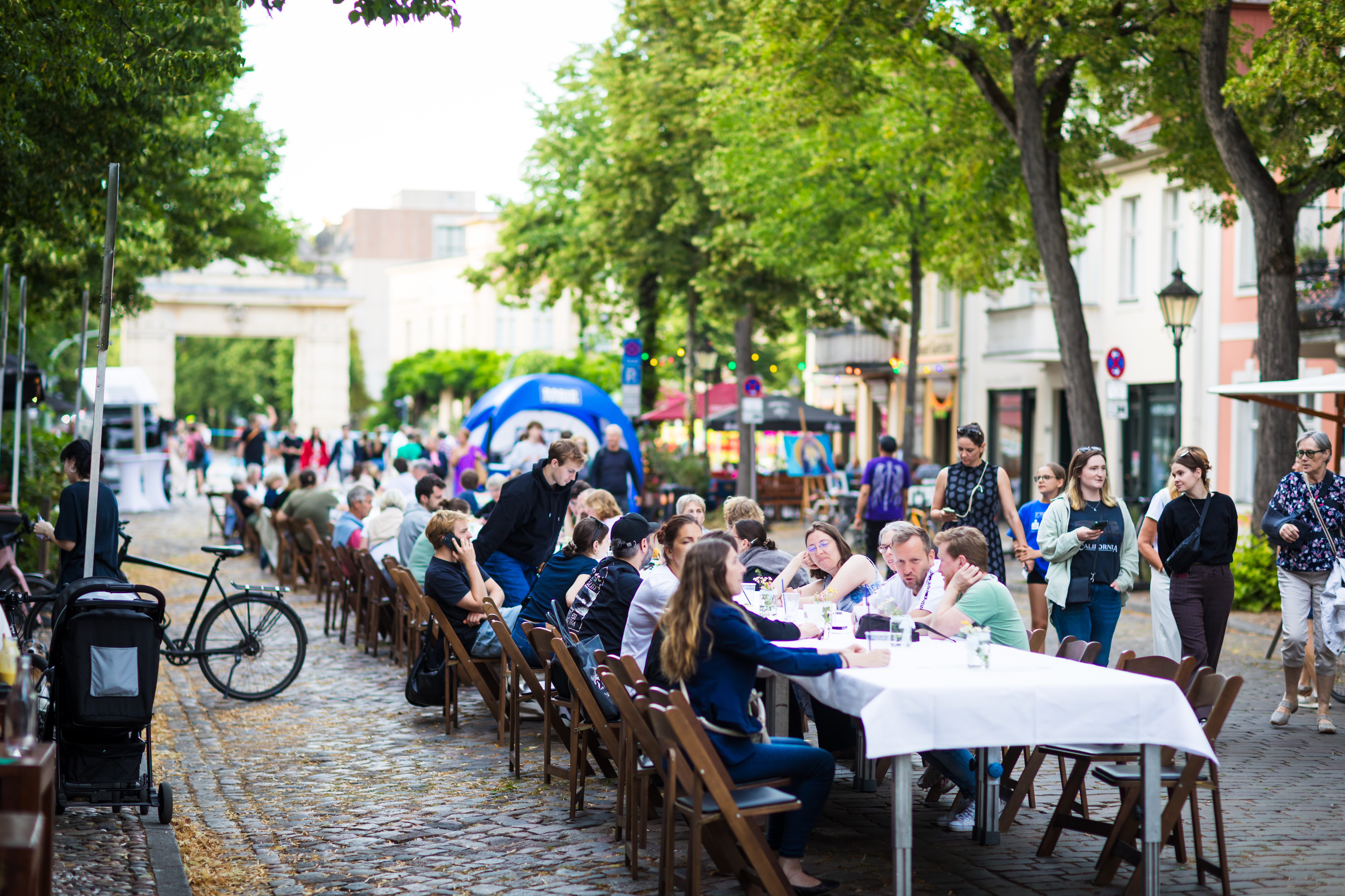 Menschen sitzen an einem langen, gedeckten Tisch in der Mitte einer Kopfsteinpflasterstraße in Potsdam und unterhalten sich.  