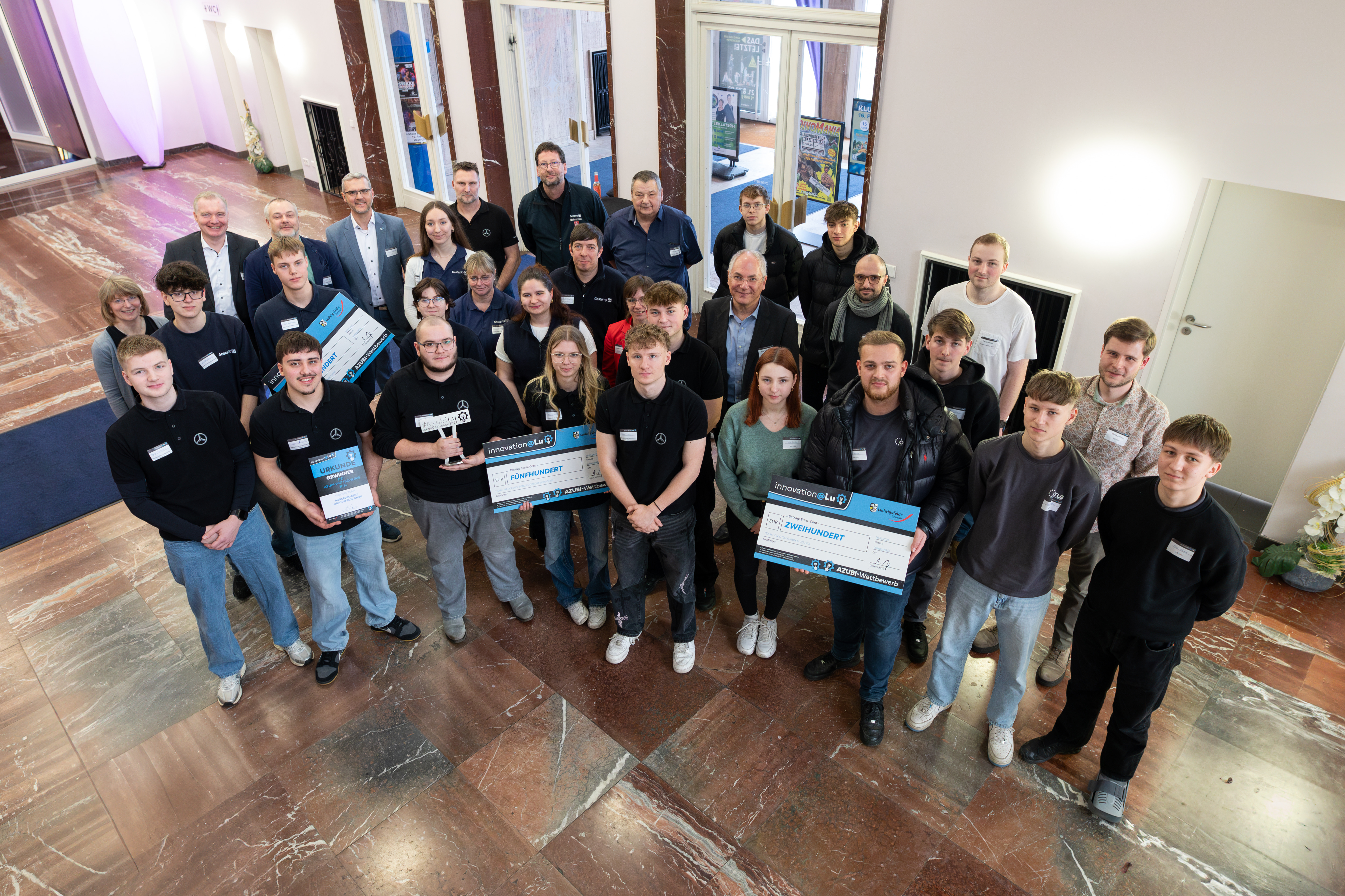 Blick aus der Luft auf eine Gruppe von Azubis in Ludwigsfelde, die für das Foto nach oben schauen und symbolische Schecks in Form von Schildern in der Hand halten.  