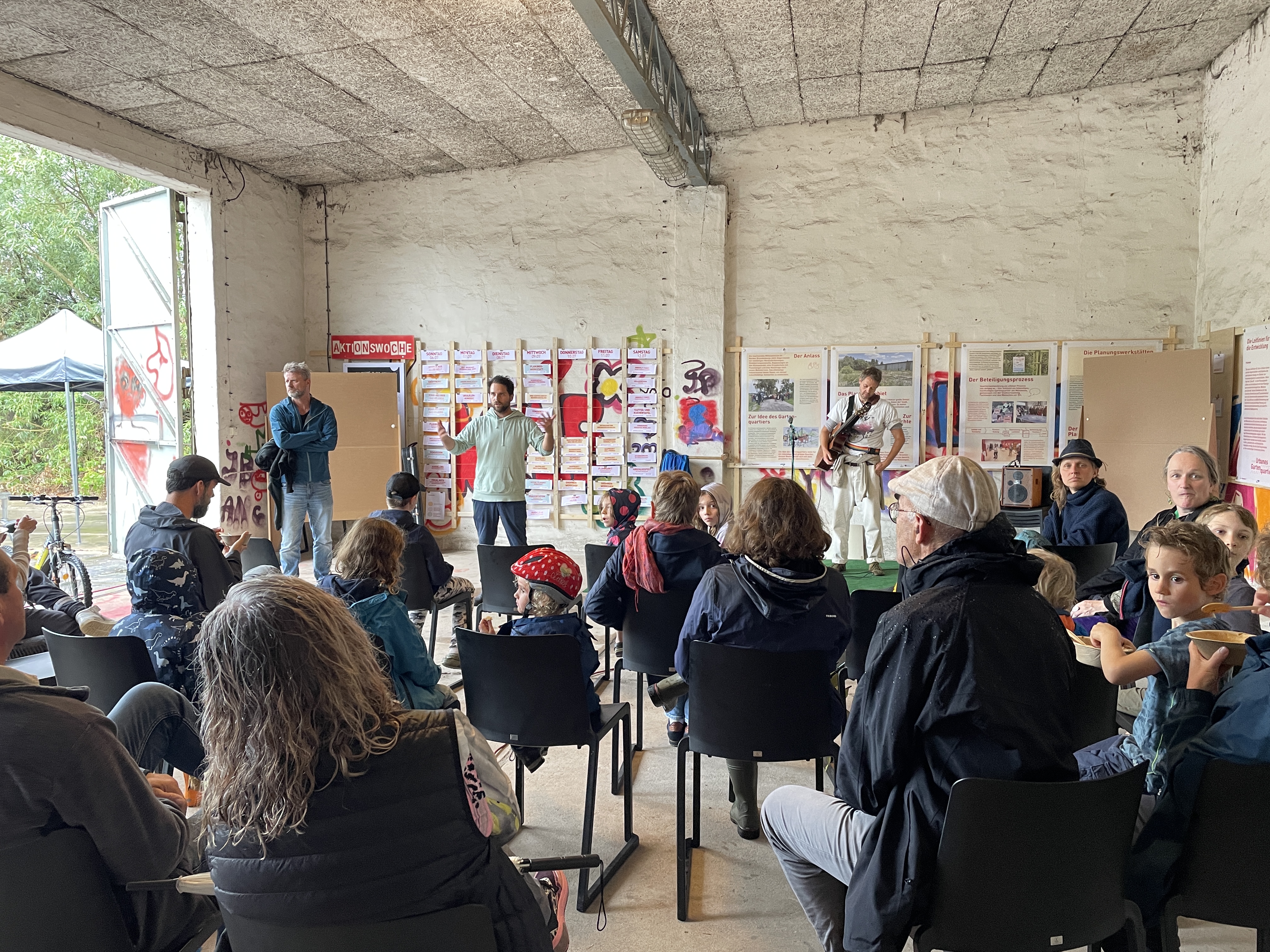 Mehrere Personen hören bei einer Präsentation vor einer Wand mit Plakaten zu. 